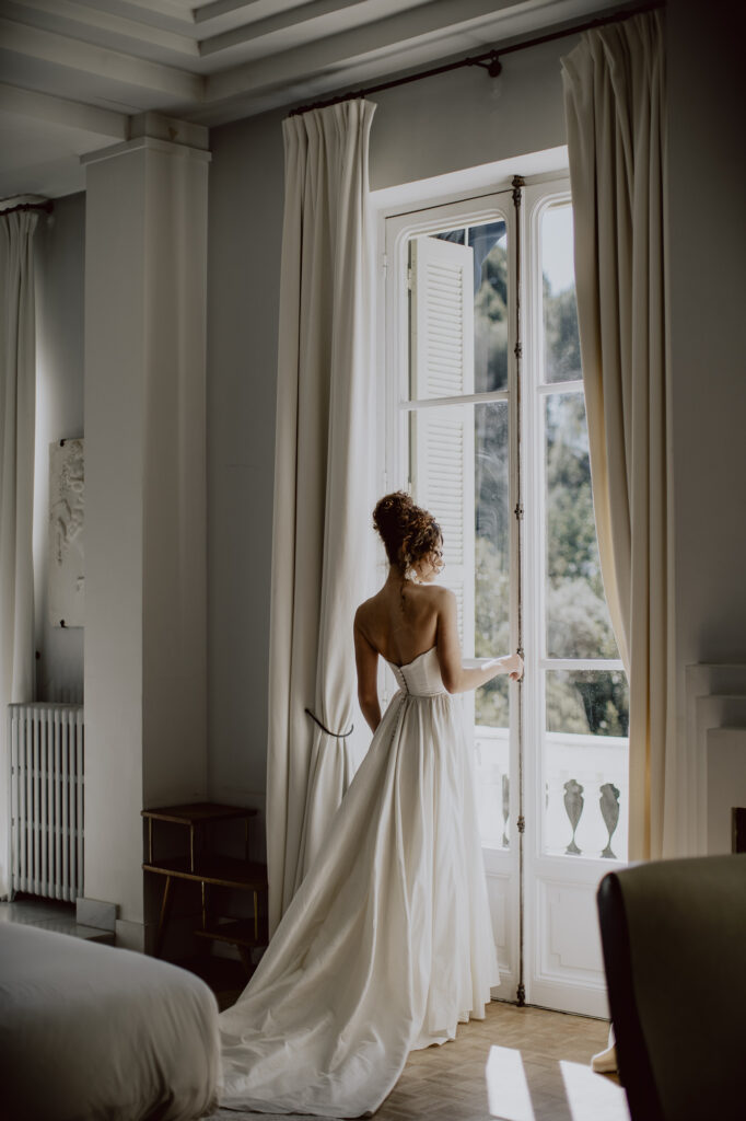 Mariée de dos en robe de mariée fluide regardant par la grande fenêtre de la Villa Rocabella face à la mer